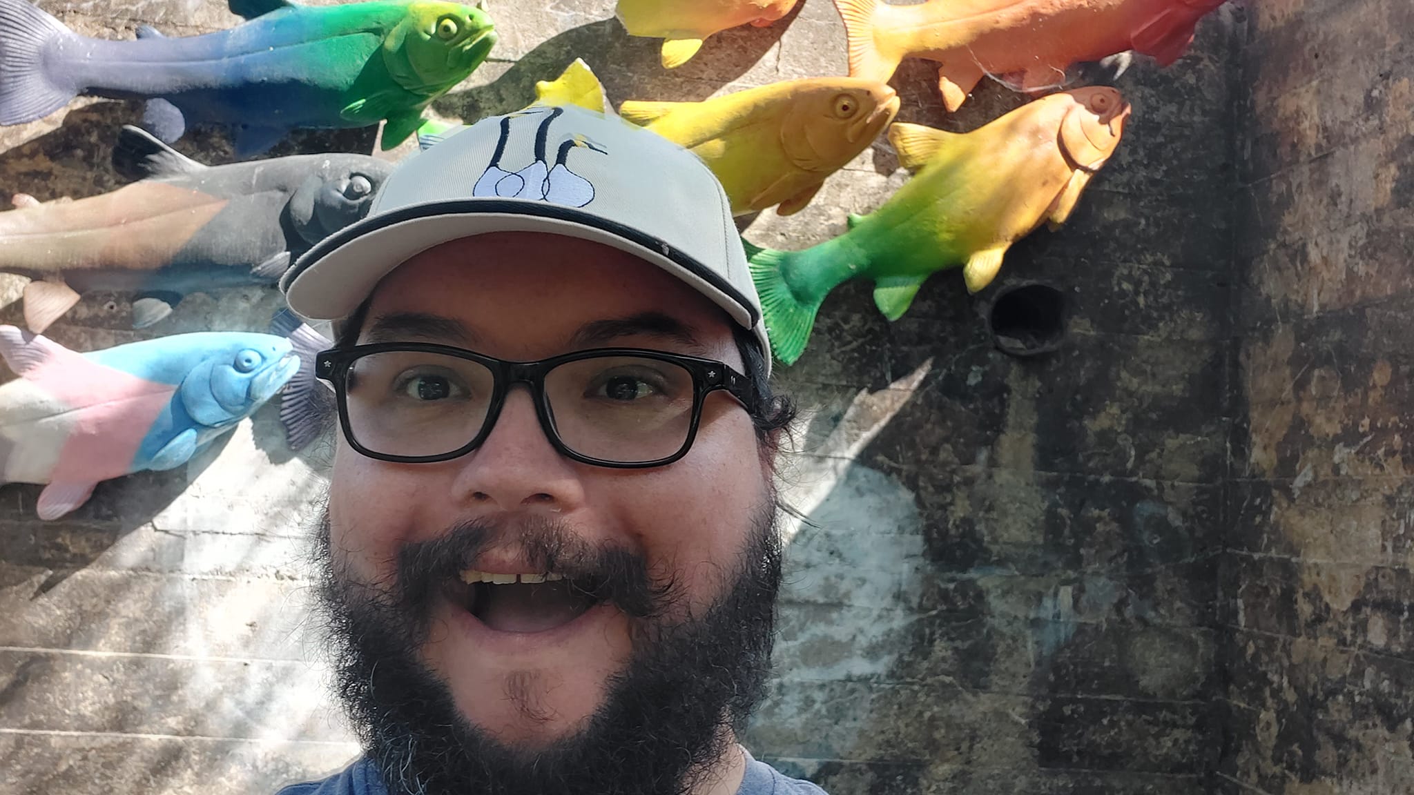 A man with short hair and bushy facial hair stands in front of some fish sculptures bolted onto a wall, they're painted rainbow colours, one is painted in trans flag colours